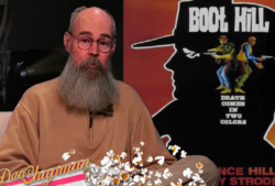 man with long beard and classes sits in front of "Boot Hill" movie poster with graphic of movie popcorn at lap level