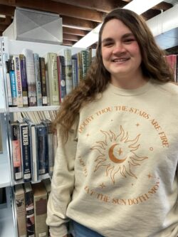 Emma wears a tan sweatshirt with a Shakespeare (Macbeth) quote in a cire around the sun and moon. She stands by the bookshelves with the Bard's works.